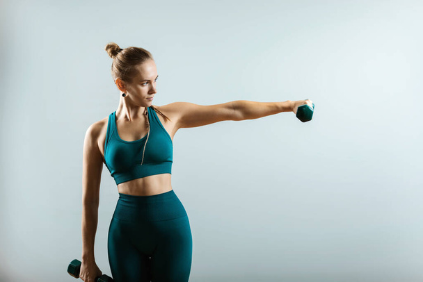 Mooi fitness model doet oefeningen met halters in handen op een grijze achtergrond. Een close-up. Een vrouw die op camera poseert met halters. Een gezonde levensstijl. Doelen bereiken, sportmotivatie. Kopieerruimte - Foto, afbeelding