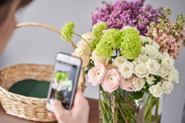 A woman takes a photo on her phone of flowers. Floral shop concept . Florist woman creates flower arrangement in a wicker basket. . Flowers delivery. - Zdjęcie, obraz