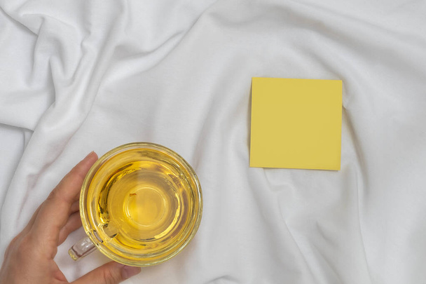 Woman hand holding cup of tea and empty sticky note on white bed background - Photo, Image