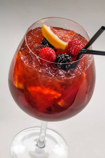 Cocktail in a transparent glass goblet, with slices of orange, raspberry,blackberry, pieces of ice and with black straws for cocktail on a light background - Foto, imagen