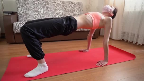 Teenager brunette slim girl in face mask in pink sport bra, black pants, white socks doing yoga poses on red mat at home against a couch. Beautiful fit woman make asana. Front view. Isolation concept. - Footage, Video