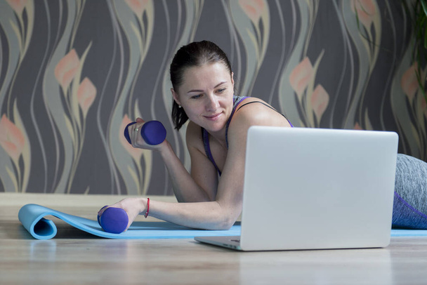 Жінка в спортивному одязі на підлозі з гантелями і використовує ноутбук вдома у вітальні. Інтернет-концепція тренування
. - Фото, зображення