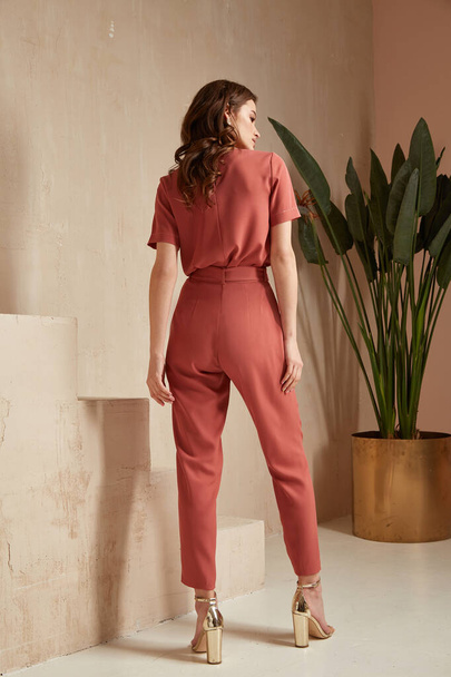 Beautiful woman fashion model brunette hair tanned skin wear pink red overalls suit sandals high heels accessory bag clothes style journey safari summer collection plant flowerpot wall stairs. - Photo, Image
