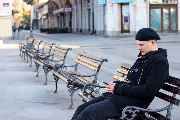  Одинокий молодой человек сидит один на скамейке на пустой площади в солнечный зимний день, пишет сообщение или ждет свидания. Концепция одиночества
. - Фото, изображение