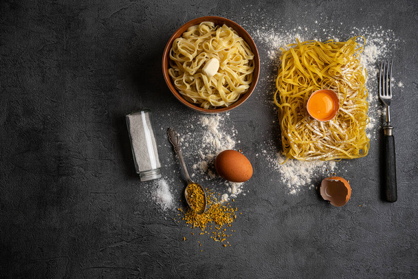Raw uncooked homemade italian pasta tagliatelle with, pasta cutter, bowls with white flour and broken egg on old wood cutting board over dark wooden background. Top view with space for text - 写真・画像
