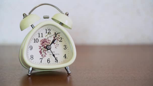 White vintage o-clock alarm clock with bells stand on a brown wooden table on blurred background close up with copy space. Classic old modern bell watch show about 1.25 p.m. local time. - Filmati, video
