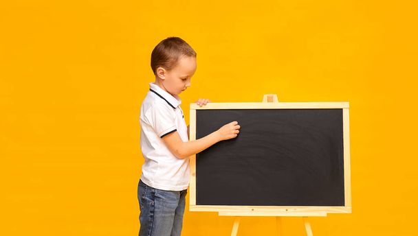 黄色の背景に白いTシャツを着た可愛い男子生徒が黒板に手を書きます。バナーだ。テキスト用の場所。学校の時間だ。家庭教育。隔離. - 写真・画像