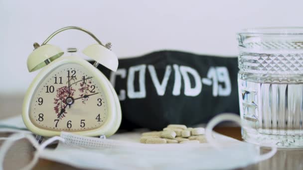 Glass with water, pills on medical face mask, black protective mask and clock on wooden table close up. White vintage alarm o-clock ringing, showing about 7.00 a.m. The concept of healing from virus. - Footage, Video