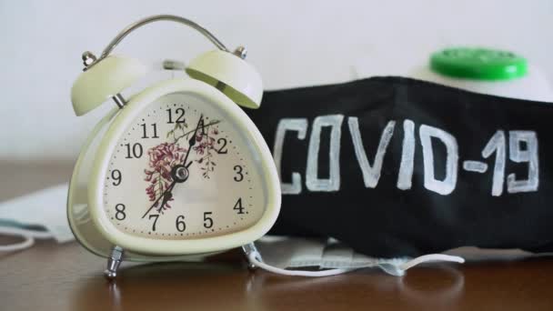 Relógio de alarme retro branco tocando na mesa de madeira marrom perto de máscaras protetoras pretas com a palavra COVID-19 de perto. O-clock mostra cerca de 7: 00 Quarentena por coronavírus covid-19 ameaça de vírus. Bloqueio
 - Filmagem, Vídeo