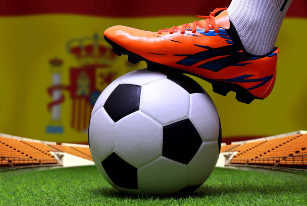Κλείστε τα πόδια και τα πόδια του ποδοσφαιριστή ή ποδοσφαιριστή με τα πόδια σε πράσινο γρασίδι έτοιμο να παίξει αγώνα σε εθνικό φόντο σημαία της Ισπανίας. - Φωτογραφία, εικόνα