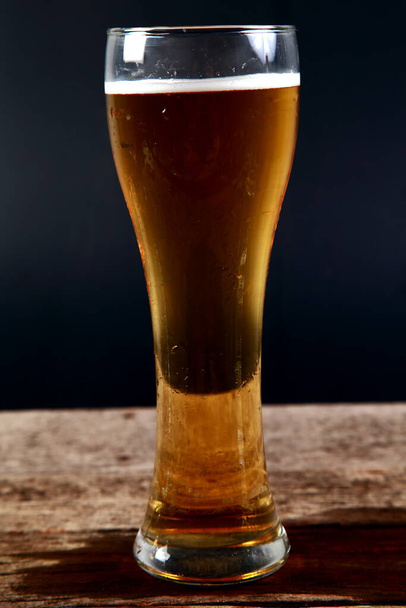 Un vaso de cerveza descansando sobre un trozo de madera con un fondo negro
. - Foto, Imagen