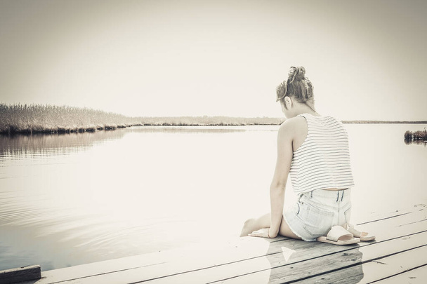 A lonely, sad girl, a teenager, on the bank of a river, a lake. The shoulders and head are down. Lack of support, understanding. Muted tone, vignetting. - Photo, Image