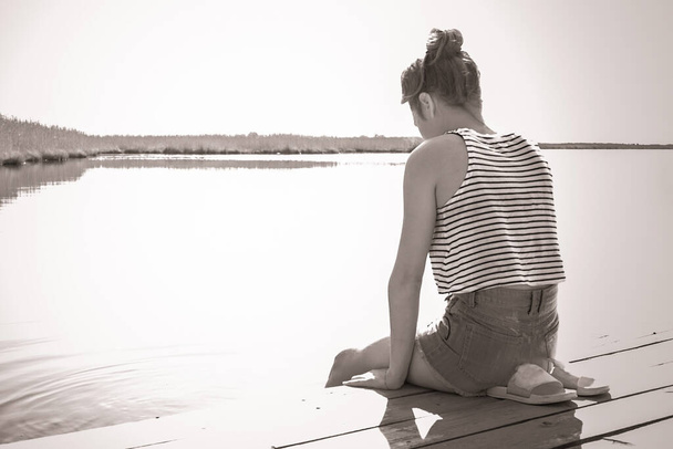 Підліток, молода дівчина на березі річки. Сумний вигляд, вислизнуті плечі. Дивиться на воду, сумно. Проблеми з підлітком, самотність. Сонячне освітлення, чорно-біле, віньєтування
. - Фото, зображення