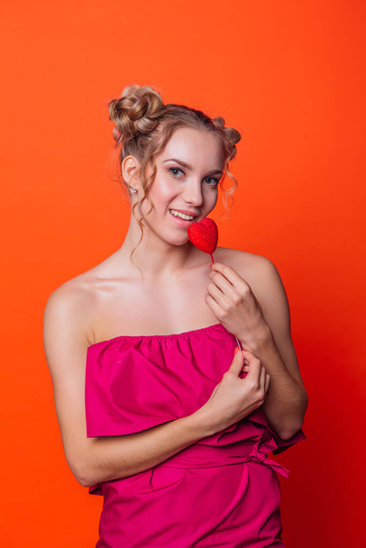 The girl in a red dress on a orange background in the studio. Blonde girl with two hair knots holding red heart on the stick. - Фото, изображение
