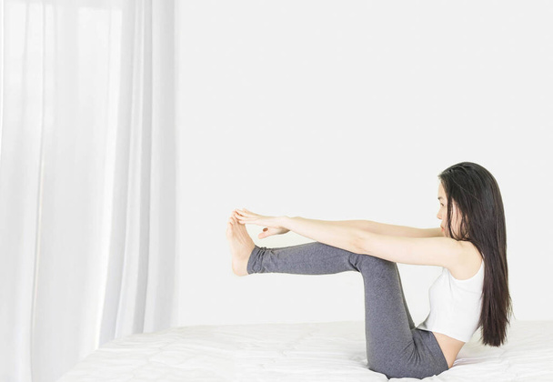 joven asiático mujer usando blanco undershirt y gris pantalones ejercicio yoga mientras sentado en un blanco cama por la ventana con un delgado cortina
, - Foto, imagen