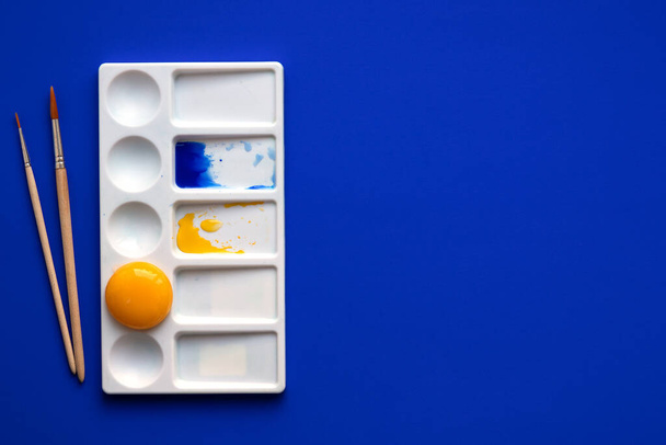 White palette with watercolor paints and brushes. Blue background. Instead of yellow, the egg yolk is used. Creative design and art. Top view and copy space. - Photo, Image
