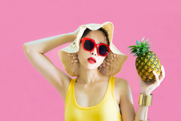 Happy beautiful asian woman in yellow undershirt,sunglasses fashion holds pineapple in her hands posing with pineapple, female funny with pineapple in summer holidays isolated on pink background - Фото, зображення