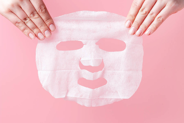Γυναικεία χέρια κατέχουν λευκή υφασμάτινη μάσκα σε ροζ φόντο. - Φωτογραφία, εικόνα