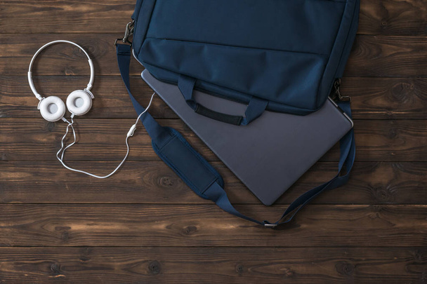 White headphones with a wire and a laptop in a blue bag on a wooden table. - Fotografie, Obrázek