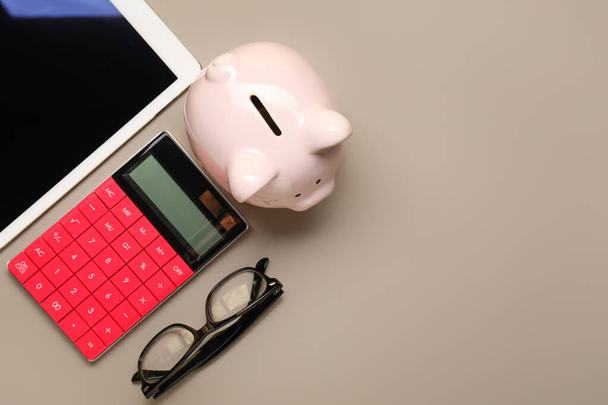 Calculadora com banco porquinho, computador tablet e óculos em fundo cinza
 - Foto, Imagem