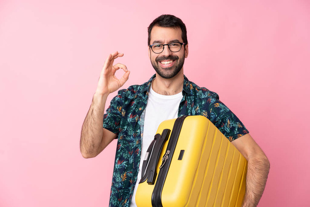 Jonge blanke man over geïsoleerde achtergrond in vakantie met reiskoffer en het maken van OK teken - Foto, afbeelding