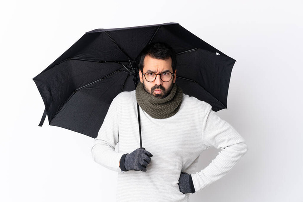 Кавказький вродливий чоловік з бородою, що тримає парасольку над ізольованою білою стіною, злий - Фото, зображення