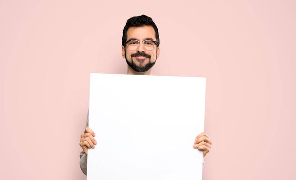 schöner Mann mit Bart, der ein leeres Plakat vor isoliertem rosa Hintergrund hält - Foto, Bild