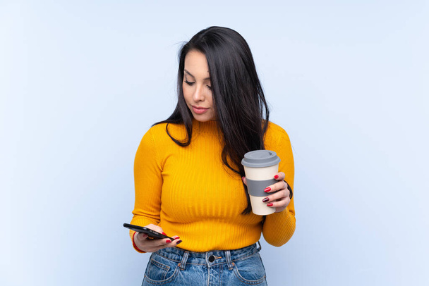 Junge Kolumbianerin vor isoliertem blauen Hintergrund mit Kaffee zum Mitnehmen und einem Handy - Foto, Bild