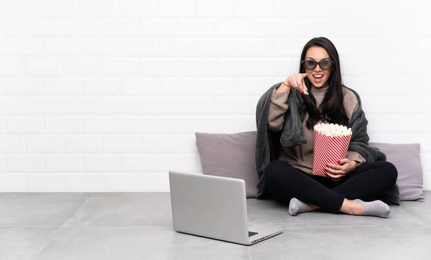 Young Colombian girl in a room with 3d glasses and holding a big bucket of popcorns while pointing front - Photo, Image