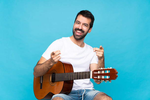 Jovem com guitarra sobre fundo azul isolado aponta dedo para você enquanto sorri
 - Foto, Imagem