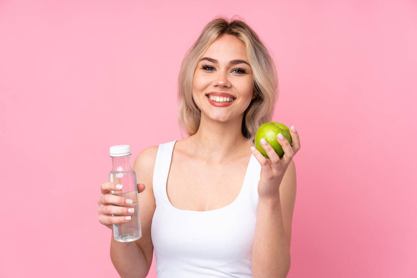 Teenager blonde girl over isolated pink background with an apple and with a bottle of water - Foto, Imagen