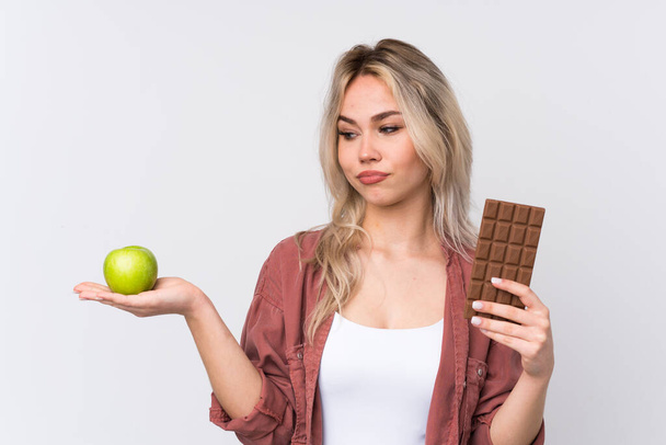 Adolescente chica rubia sobre fondo blanco aislado que tiene dudas mientras toma una tableta de chocolate en una mano y una manzana en la otra - Foto, imagen
