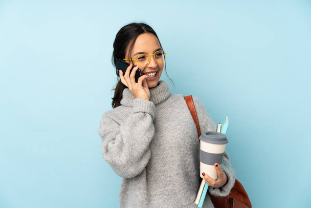 Jonge gemengde ras vrouw gaat naar school geïsoleerd op blauwe achtergrond met koffie mee te nemen en een mobiele - Foto, afbeelding