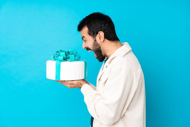 Молодой красивый мужчина с большим тортом на изолированном синем фоне - Фото, изображение