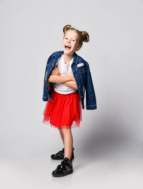 Blonde kid girl with funny buns in red skirt, white t-shirt and denim jacket over shoulder poses having fun  - Photo, image