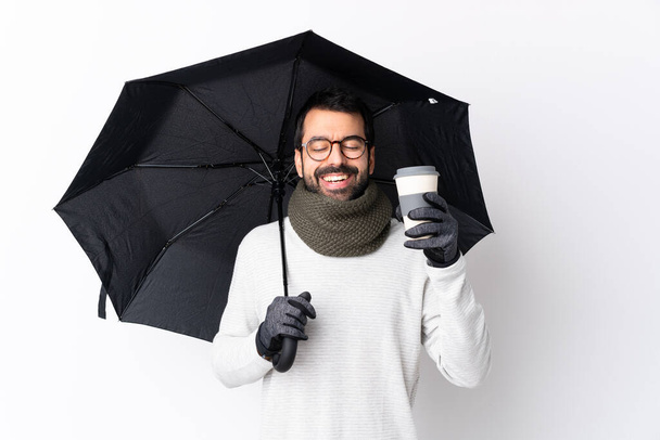 Biały przystojniak z brodą trzymający parasol i kawę do zabrania przez odizolowaną białą ścianę - Zdjęcie, obraz