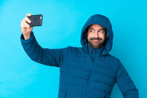Hombre vistiendo chaqueta de invierno y sosteniendo un café para llevar sobre fondo azul aislado haciendo una selfie
 - Foto, imagen