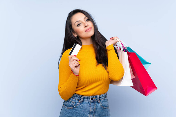 Young Colombian girl over isolated blue background holding shopping bags and a credit card - Photo, image