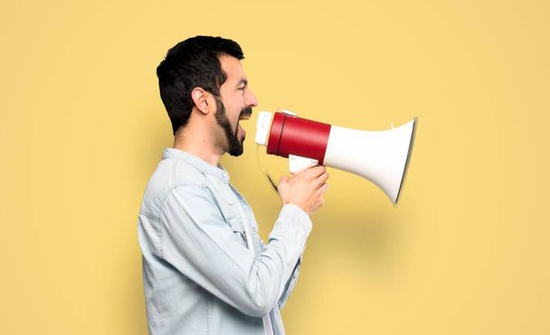 Handsome man with beard shouting through a megaphone over isolated yellow background - Foto, Bild