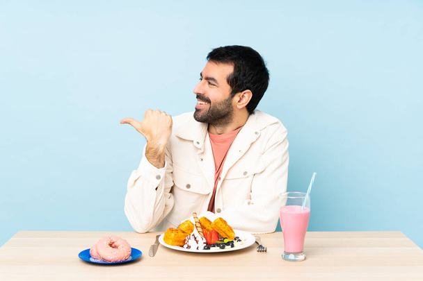 Ο άνθρωπος σε ένα τραπέζι έχοντας βάφλες πρωινό και ένα milkshake δείχνοντας προς τα πλάγια για να παρουσιάσει ένα προϊόν - Φωτογραφία, εικόνα