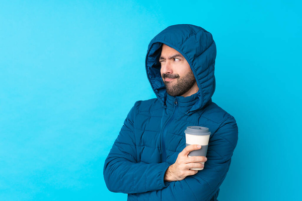 Uomo indossando giacca invernale e tenendo un caffè da asporto su isolato ritratto sfondo blu
 - Foto, immagini