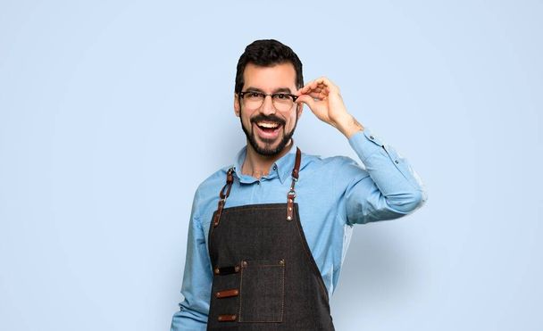 Man with apron with glasses and surprised over isolated blue background - Photo, image