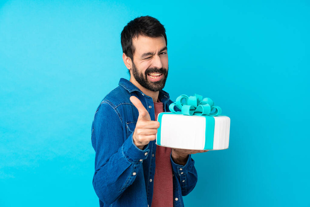 Junger gutaussehender Mann mit einer großen Torte vor isoliertem blauen Hintergrund, der nach vorne zeigt und lächelt - Foto, Bild