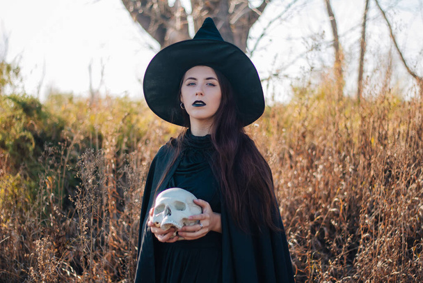 A young witch with pale skin and black lips in a black hat, a dress and a raincoat. Holds the skull of a dead person. Against the background of a tree of gallows. halloween, magic, fantasy, necromancy. - Φωτογραφία, εικόνα