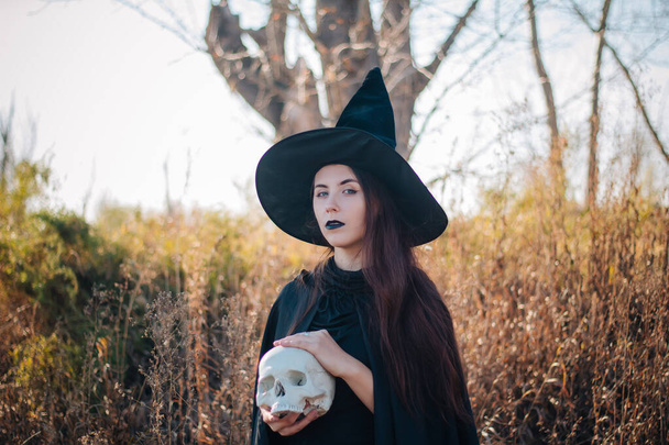Egy fiatal boszorkány sápadt bőrrel és fekete ajkakkal fekete kalapban, ruhában és esőkabátban. Egy halott ember koponyáját fogja. A háttérben egy bitófával. halloween, varázslat, fantázia, nekromancia. - Fotó, kép