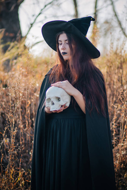 Uma jovem bruxa com pele pálida e lábios pretos em um chapéu preto, um vestido e uma capa de chuva. Segura o crânio de uma pessoa morta. Contra o fundo de uma árvore de forca. Halloween, magia, fantasia, necromancia
. - Foto, Imagem