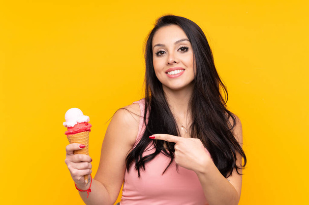 Young Colombian girl holding an cornet ice cream over isolated yellow background and pointing it - Foto, afbeelding