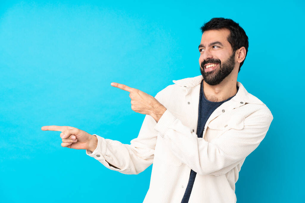 Молодой красивый мужчина в белой вельветовой куртке на изолированном синем фоне, указывая пальцем в сторону и представляя продукт
 - Фото, изображение