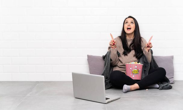 Junges kolumbianisches Mädchen hält eine Schale Popcorn in der Hand und zeigt überrascht einen Film in einem Laptop - Foto, Bild