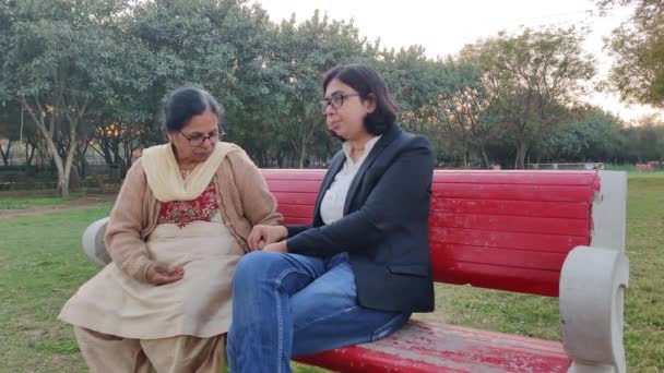 Strzał starszej emerytowanej Hinduski siedzącej w parku z synową na czerwonej ławce obierającej orzeszki i jedzącej je. Koncepcja - szczęśliwe emerytury  - Materiał filmowy, wideo